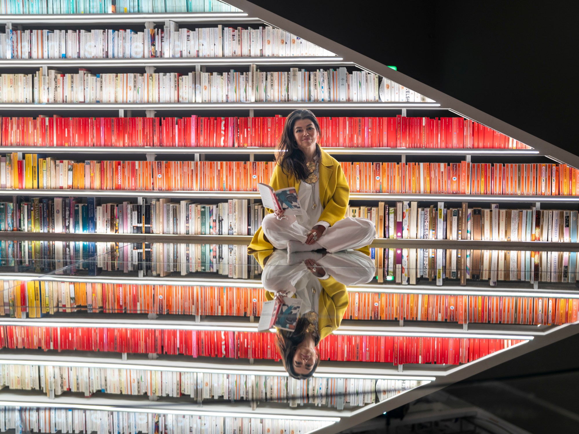 Es Devlin sits on a mirrored floor in front of the rainbow-ordered shelves of her Infinite Library at the Design Museum