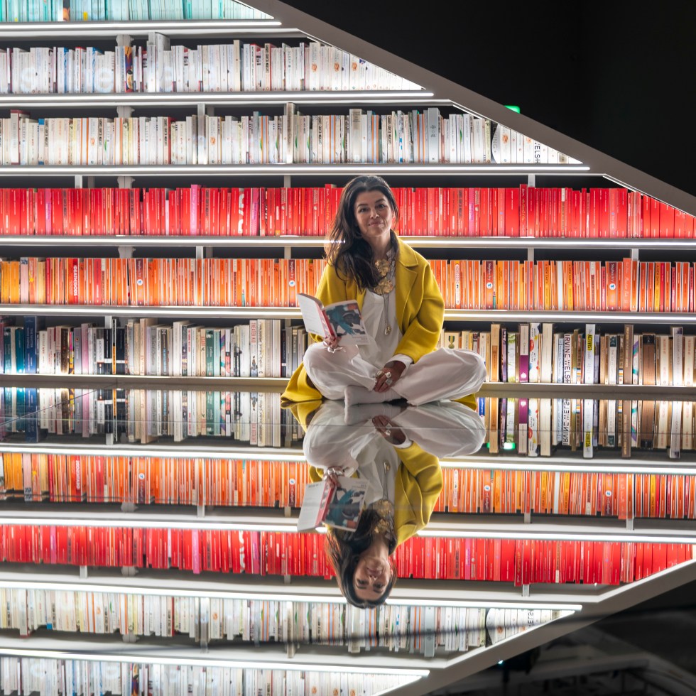 Es Devlin sits on a mirrored floor in front of the rainbow-ordered shelves of her Infinite Library at the Design Museum